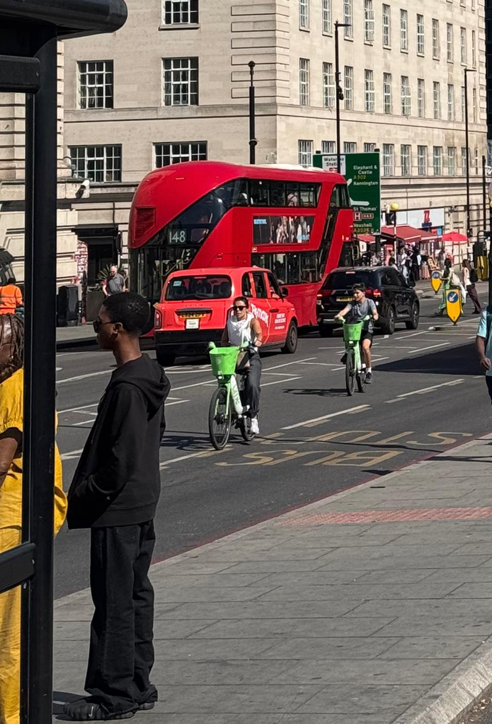 A modern London street scene on a sunny day, featuring a bright red New Routemaster double-decker bus, a cyclist on a green rental bike, and pedestrians waiting to cross the road.