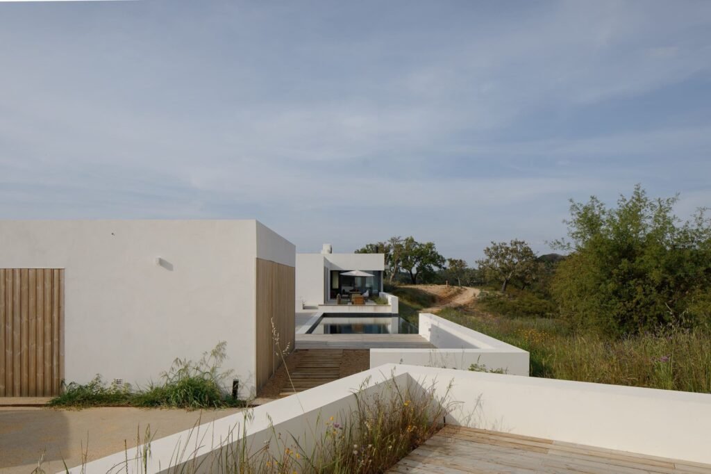 Exterior view of Melides MG House showing white detached volumes with wooden panels, outdoor deck, and swimming pool leading toward the landscape.