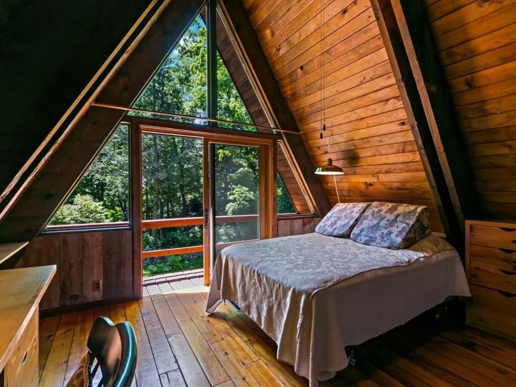 Another upper-level A-frame bedroom with vaulted, wood-paneled walls. Glass doors lead to a balcony with views of trees. The bed has a unique, high floral-patterned backrest. A simple modern lamp hangs from the ceiling. A leather lounge chair is partially visible on the bottom left.