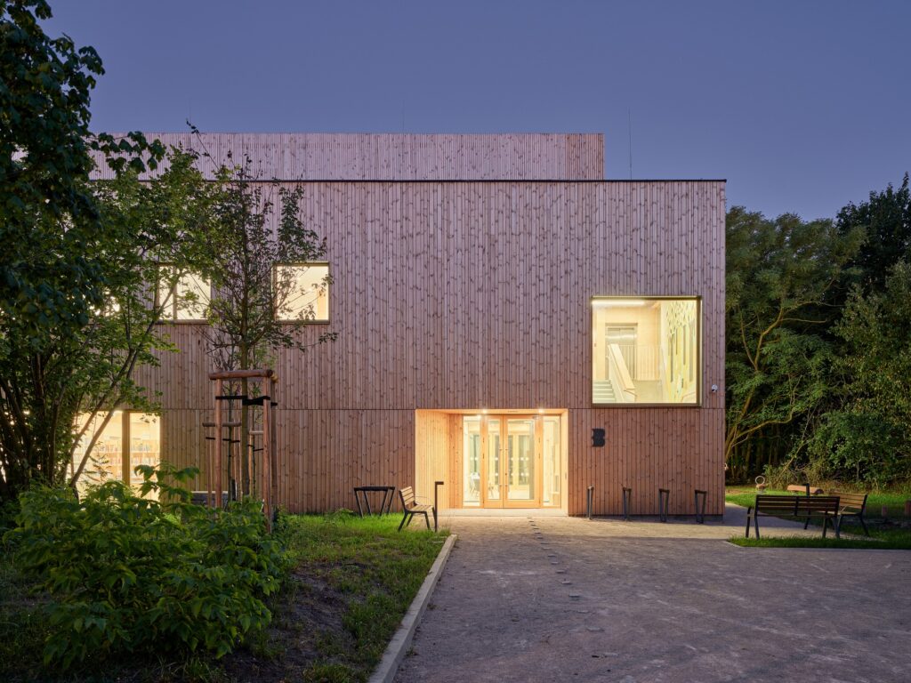 Evening view of the illuminated wooden facade and main entrance of the Trzeboszka Public Library showcasing interior warmth.