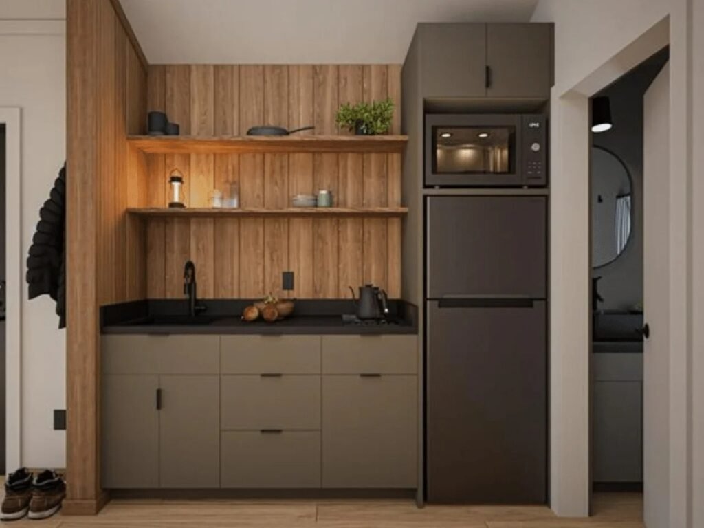 Compact modern kitchenette in the A-Frame Bunkhouse with taupe cabinets, dark countertop, open timber shelving, built-in microwave, full-size refrigerator, and matte black fixture.