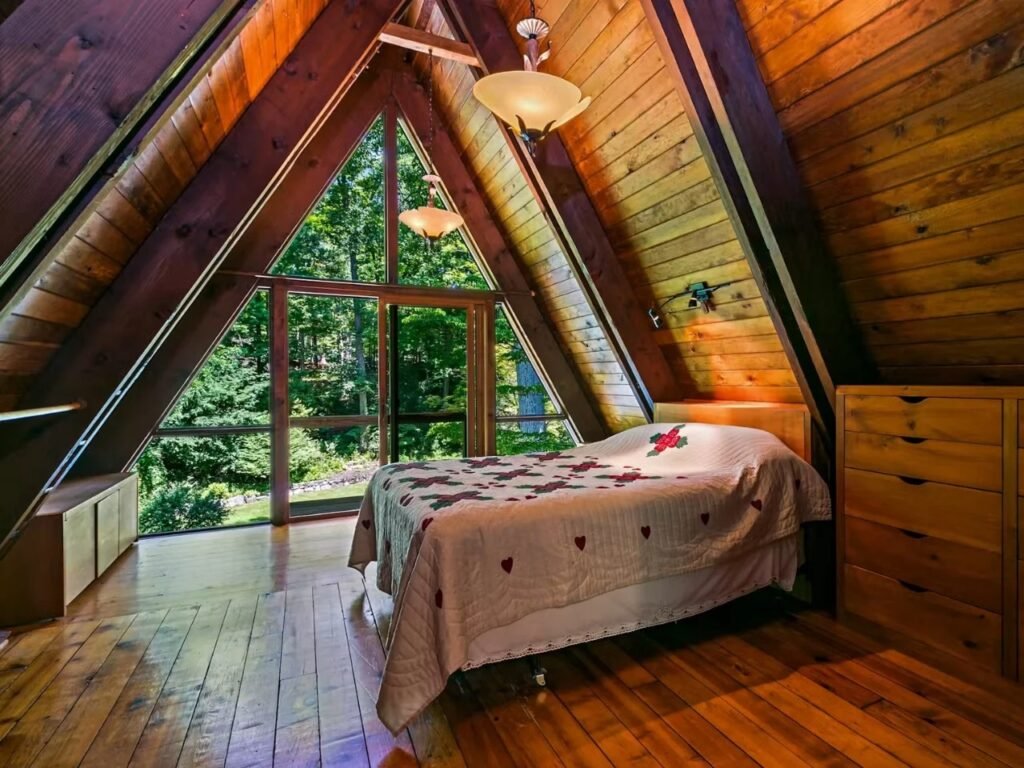 A bedroom on the lower level, showcasing the A-frame's steep, angular wood ceiling. Exposed beams are left as dark wood. Glass doors and windows open to a patio view of trees. A bed with a distinctive red heart-patterned quilt is centered under two contemporary pendant lights. Hardwood floors and a built-in wooden dresser are visible.
