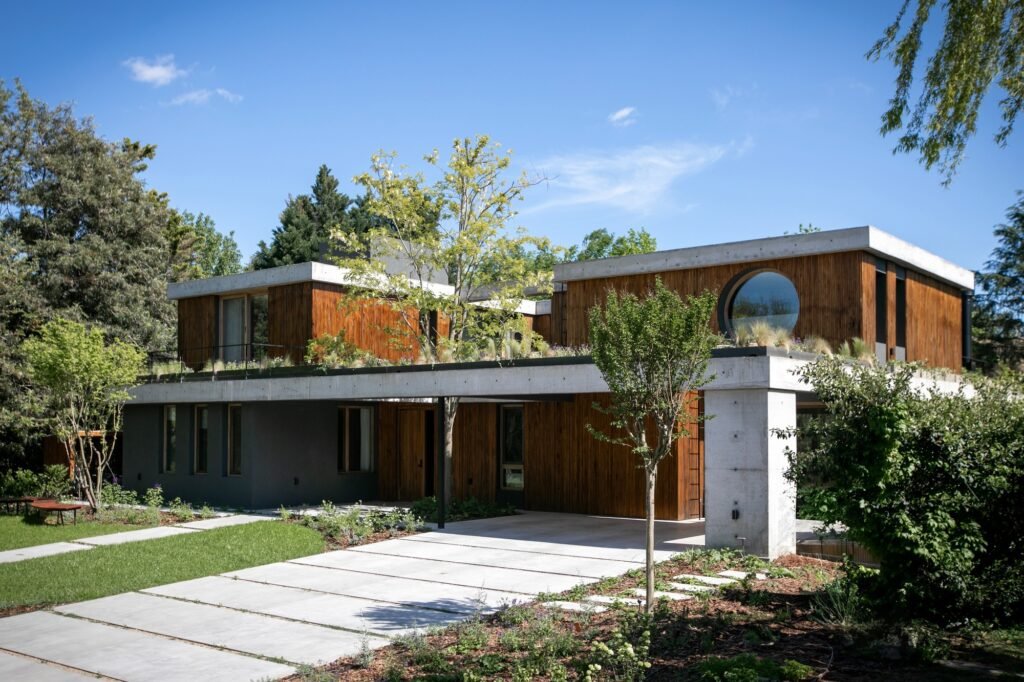 Exterior front facade of Casa MDL featuring exposed concrete slabs, vertical wood cladding, a circular window, and integrated garden landscaping.