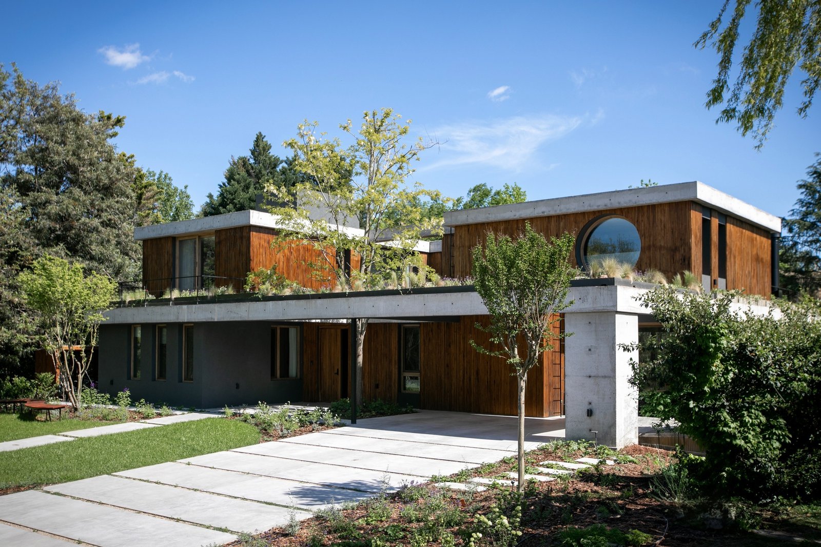 Exterior front facade of Casa MDL featuring exposed concrete slabs, vertical wood cladding, a circular window, and integrated garden landscaping.