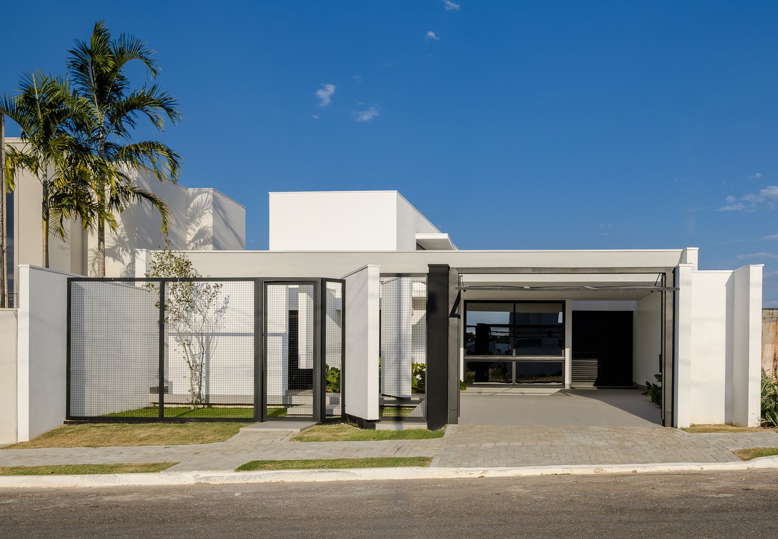 Front street view of the modern, single-story Sorriso House with white geometric volumes, a black steel grid fence, palm trees, and an open garage on a deep plot.
