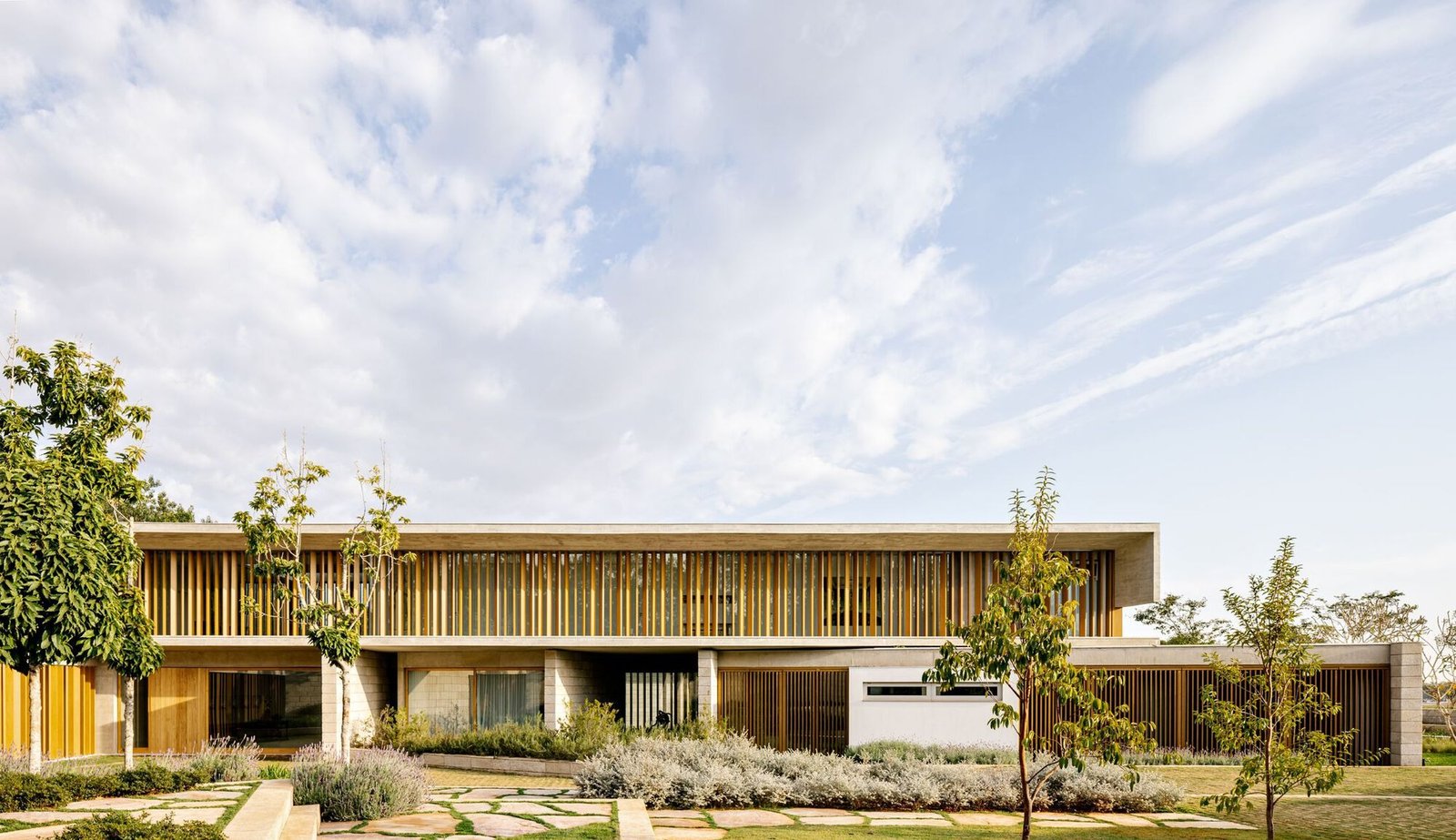 Horizontal modern house facade at Fazenda Boa Vista featuring exposed concrete, vertical wooden louvers, stone walls, and surrounding mature trees.