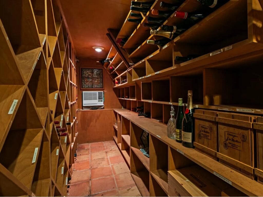 A functional wine cellar within the Woods Home, located in a lower-level space. Floor-to-ceiling wooden wine racks with labels are built into the red-tiled space. A window AC unit is visible on the far wall. Wooden crates of wine and loose bottles are stored.