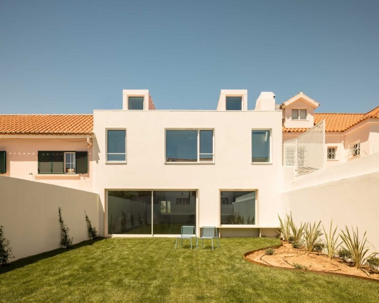 Rear elevation of a renovated 1950s house in Restelo showing large glass sliding doors, minimalist white plaster facade, and a green backyard garden.