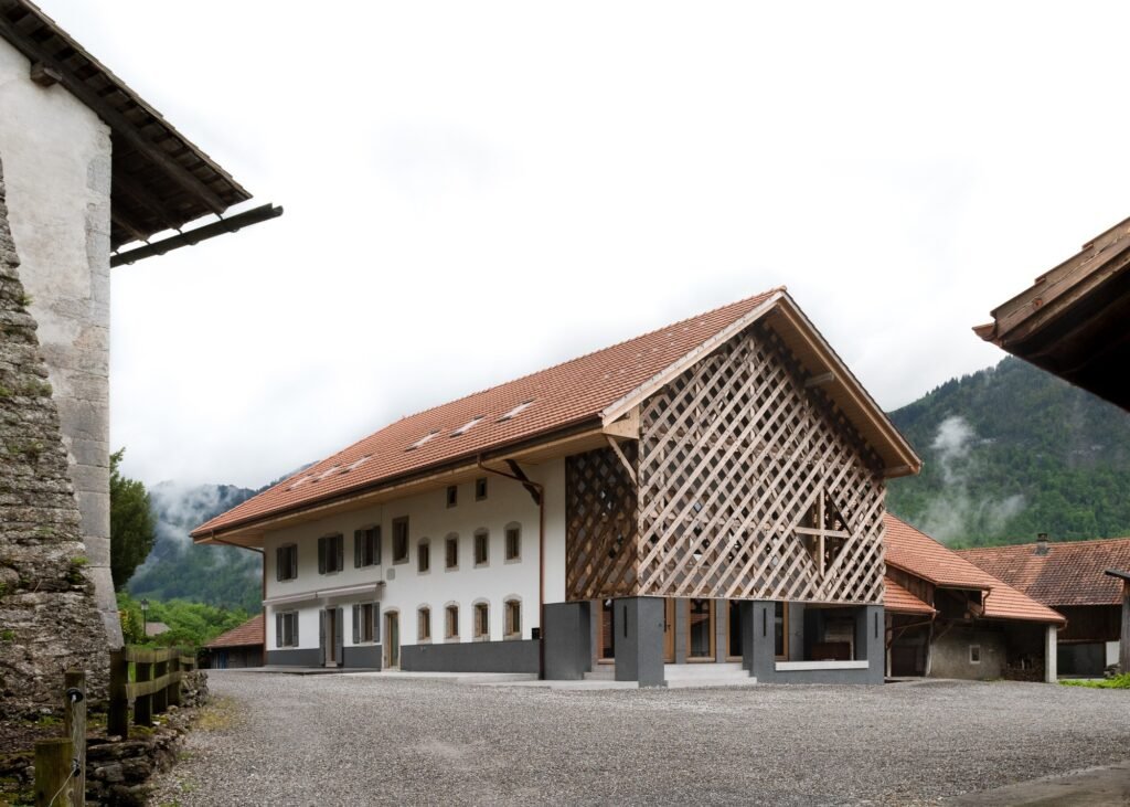 Exterior view of the renovated 18th-century Grandvillard farmhouse featuring a traditional tiled roof, white masonry facade, and an intricate wooden lattice screen on the converted agricultural wing.