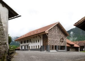 Exterior view of the renovated 18th-century Grandvillard farmhouse featuring a traditional tiled roof, white masonry facade, and an intricate wooden lattice screen on the converted agricultural wing.