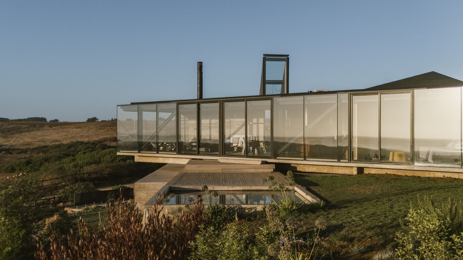 Side view of Casa 22 featuring expansive floor-to-ceiling glass facades overlooking a grassy coastal slope and a pool deck at sunset.