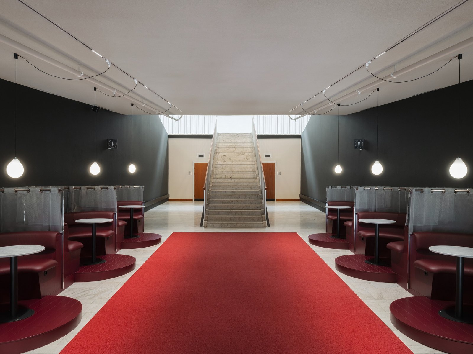 Symmetrical view of the rehabilitated main hall in Folkets Hus showing a central marble staircase flanked by red leather seating booths and a prominent red carpet.