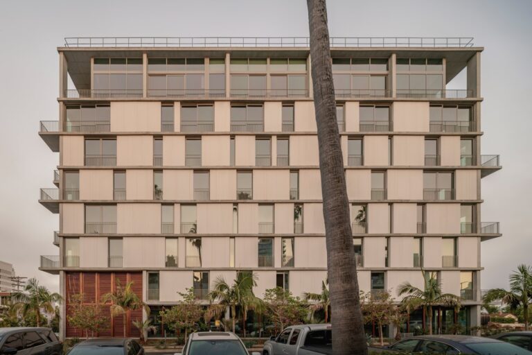 Front elevation of the KAYA building in San Diego showing exposed concrete, fiber-cement panels, and floor-to-ceiling windows framed by palm trees.
