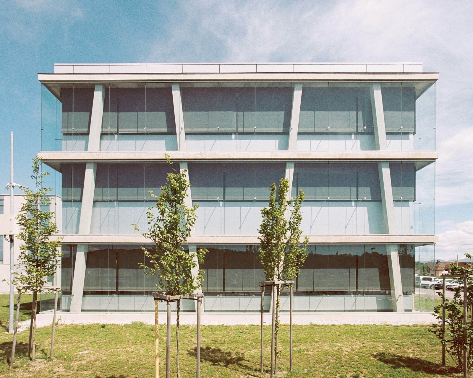 Symmetrical view of the three-story Educational Center in Ljubljana showing inclined reinforced concrete columns and double-skin glass curtain walls set against a green lawn.