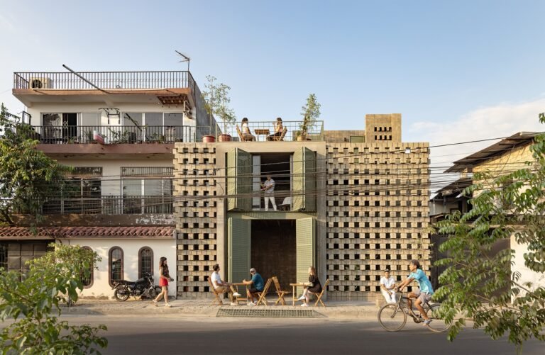Ground-level perspective of La Colmena's multi-family building in Durán, Ecuador, featuring a porous facade made of locally sourced concrete blocks, open transitional doorways, and street-level social interaction with residents sitting outside on the sidewalk and a cyclist passing.