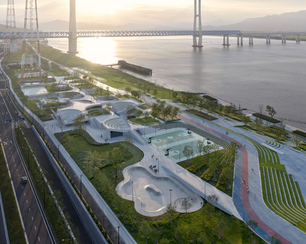 High-angle aerial view of the undulating green park along the Jiaojiang River, featuring sports courts, a concrete skate park, and the Hangzhou-Taizhou high-speed railway bridge in the background.