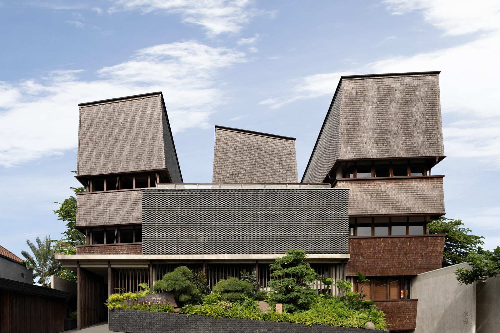 Front facade of Kemilu Ubud resort featuring reinterpreted Limasan wooden roof structures and natural stone finishes integrated with tropical greenery.