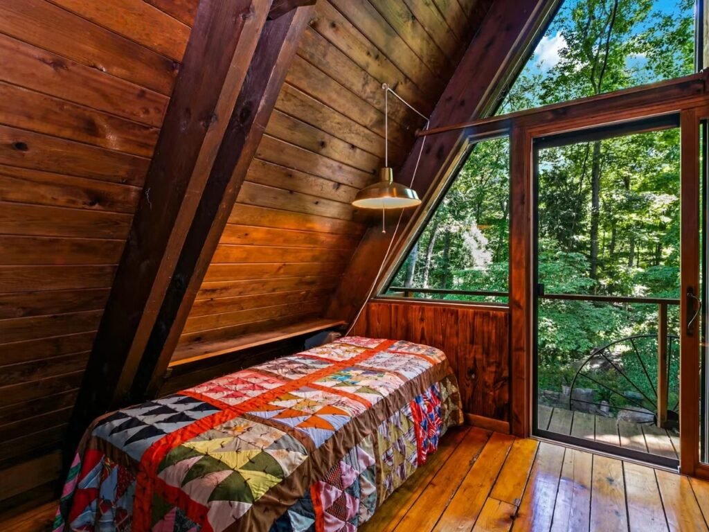 A cozy upper-level bedroom tucked under the sloping wood-paneled A-frame roof. The bed is covered with a colorful patchwork quilt, positioned next to a sliding glass door that leads to a wooden deck surrounded by vibrant green forest foliage.