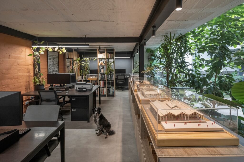 Long open-plan architectural workspace featuring display cases with physical scale models, exposed brick walls, and concrete ceiling.
