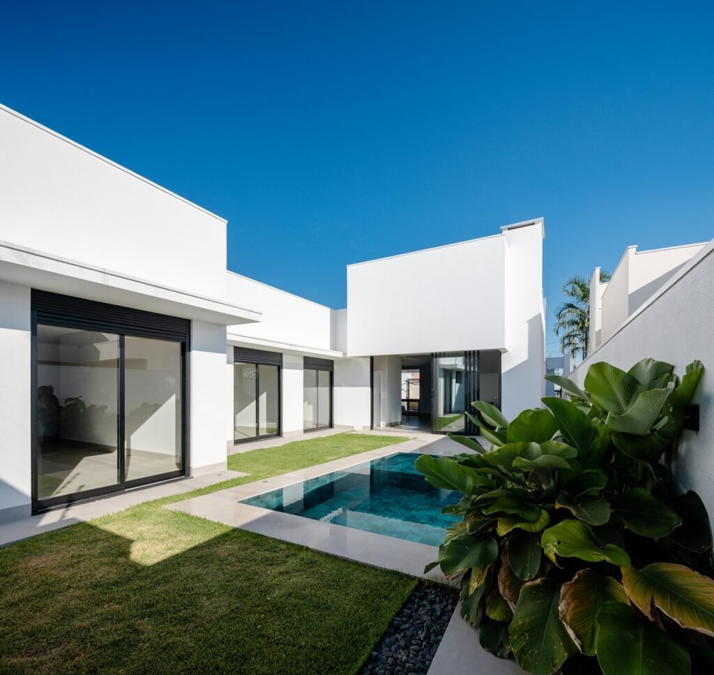 Wide-angle view of the private courtyard at Sorriso House featuring an L-shaped white building, a small rectangular swimming pool, a grassy lawn, and lush tropical foliage under a clear blue sky.