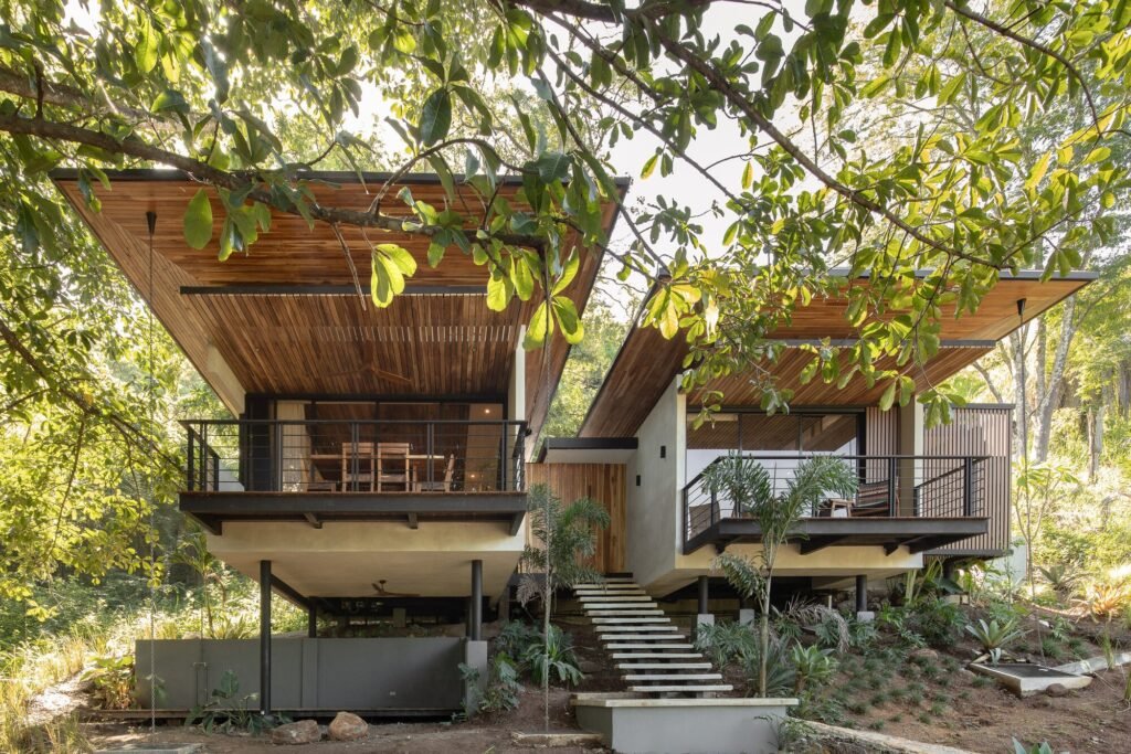 Frontal view of Villas Ra&iacute;ces featuring elevated dual volumes connected by central floating stairs on a sloped site.