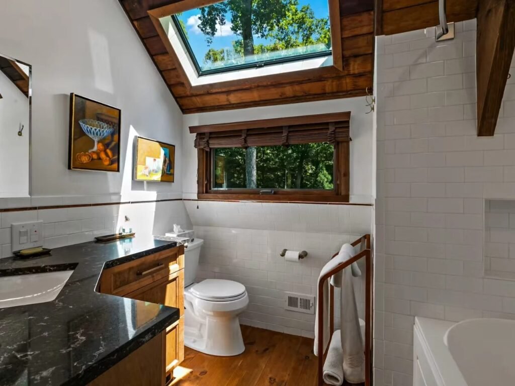A modern bathroom with white subway tile walls, a dark granite countertop, dark wood vanity, hardwood flooring, a horizontal window overlooking trees, and a large skylight set into the wood-paneled slanted ceiling.