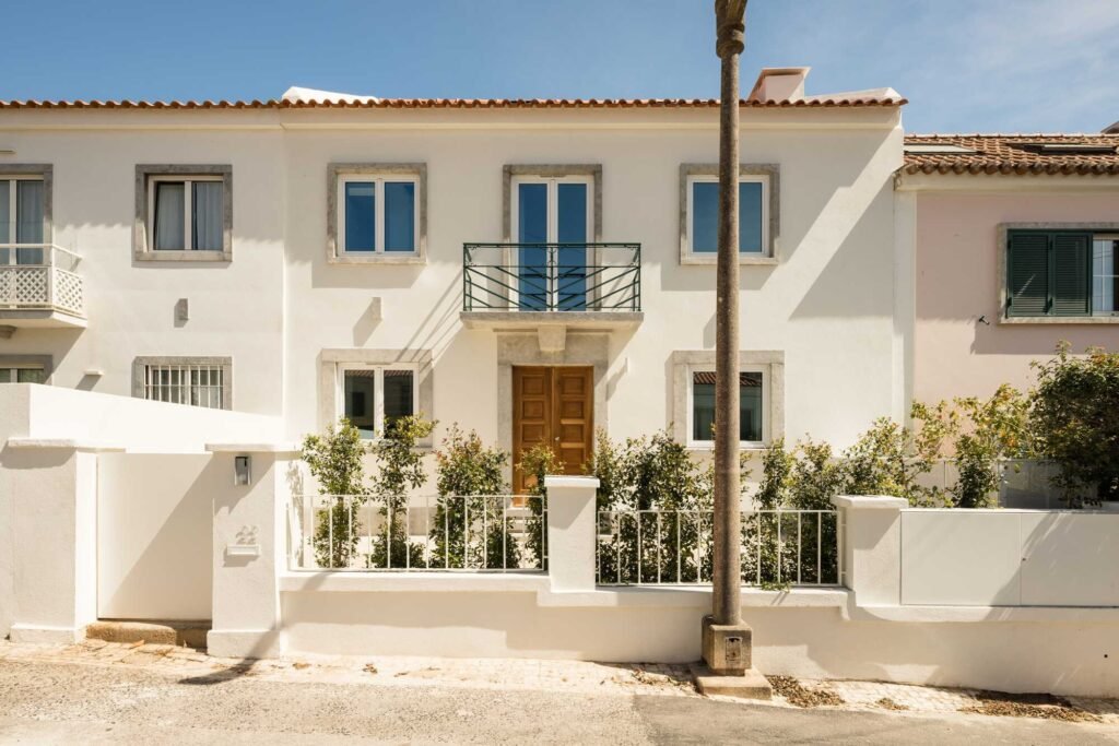 Traditional 1950s white residential street facade in Restelo with restored stone-framed windows, a central wooden door, and a black wrought-iron balcony.