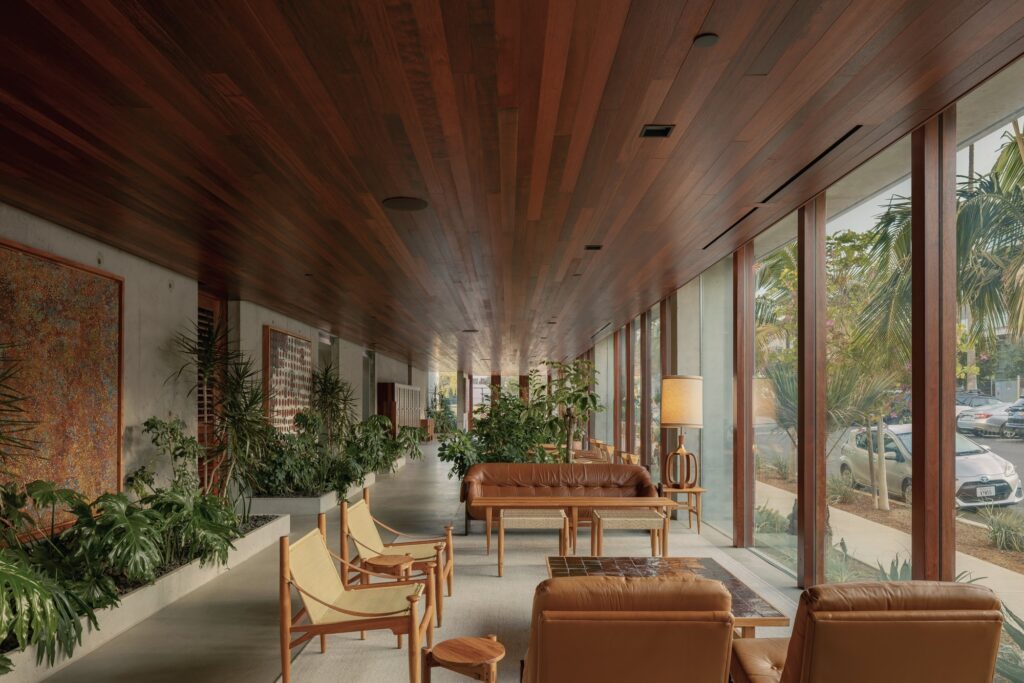 Ground-floor interior lobby of KAYA featuring rich wood ceilings, mid-century furniture, exposed concrete walls, and large glass windows.