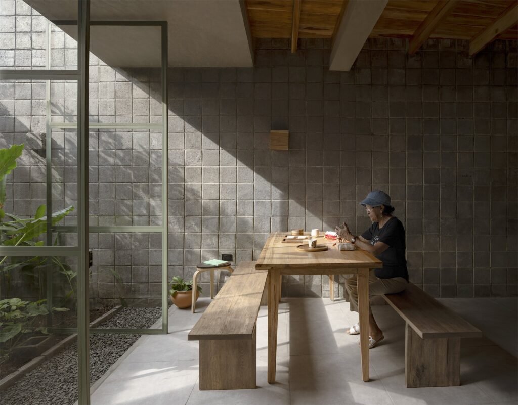 An elderly woman sitting at a long wooden communal dining table inside La Colmena, illuminated by natural sunlight filtering through a concrete block wall and an adjacent indoor patio garden.