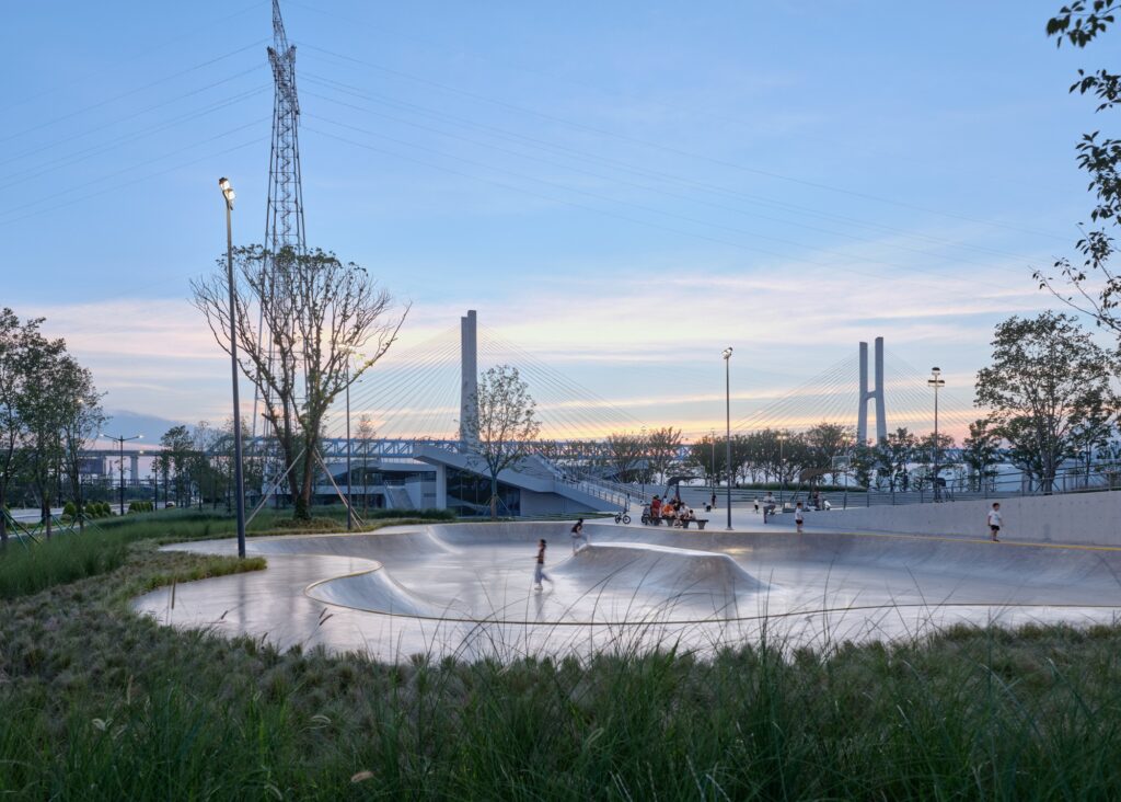 Smooth concrete skate bowl integrated into grass hills at dusk, with children playing and high-voltage transmission towers framing the cable-stayed bridge in the background.
