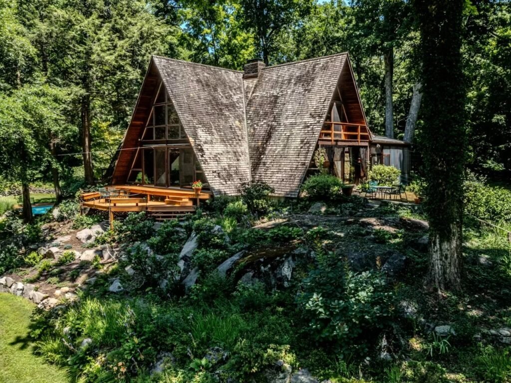 An exterior view showing the full A-frame structure elevated above natural rock outcrops, surrounded by lush green trees, featuring wide wooden decks and large glazed gables under a shingled roof.