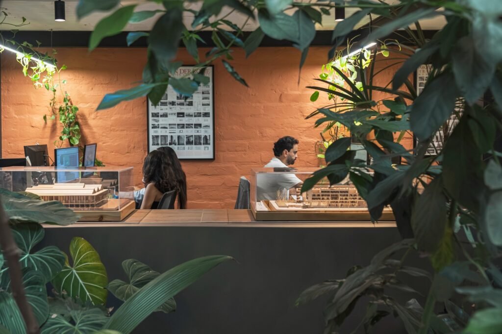 Architects working at desks behind glass model display counters framed by indoor tropical foliage and warm brick walls.