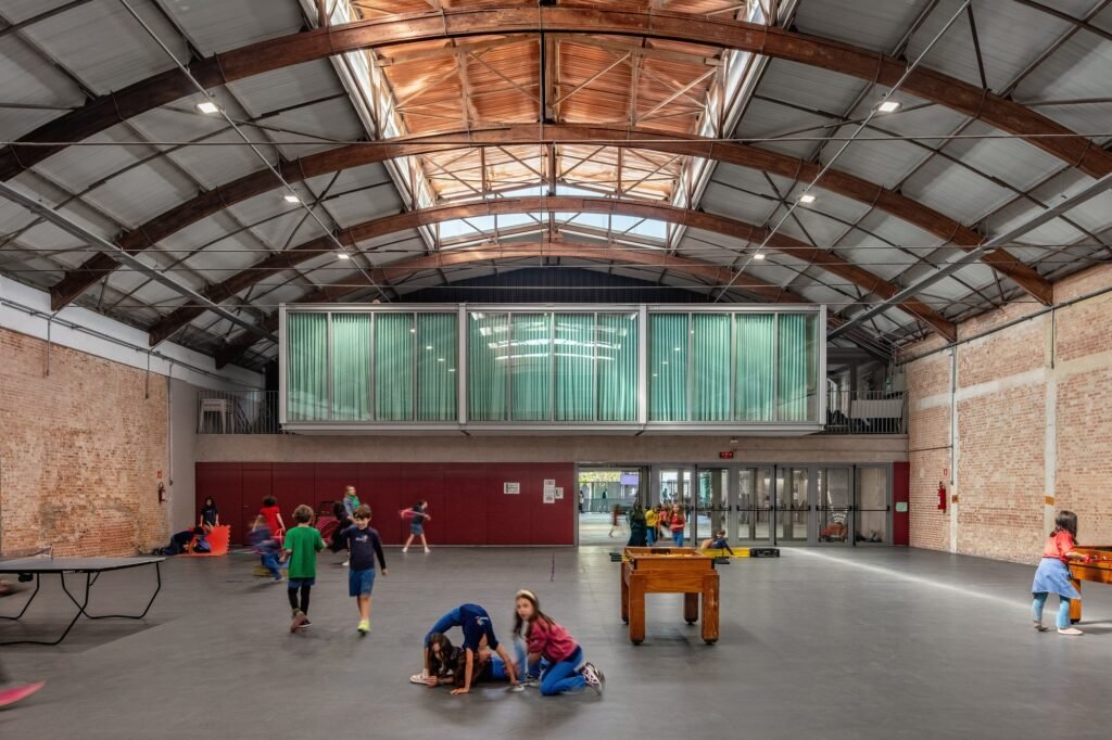 Students playing inside an adaptive reuse industrial warehouse with exposed brick walls, timber roof trusses, and elevated glass pavilion.