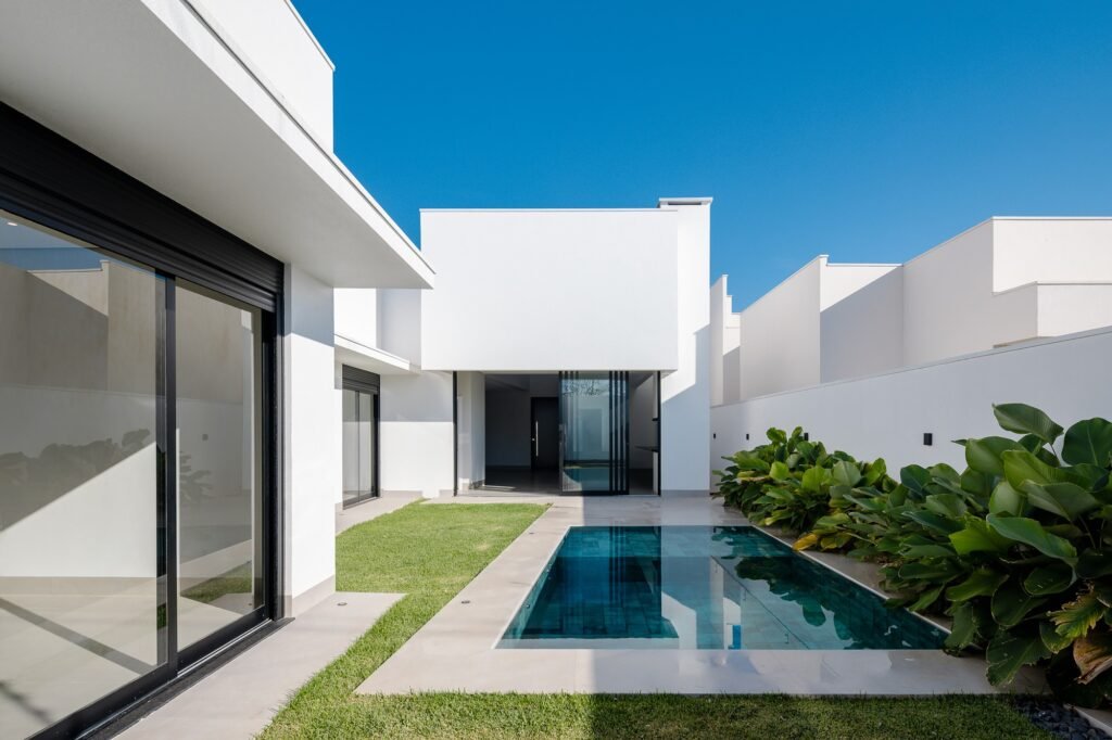 Exterior view of a longitudinal swimming pool flanked by a grassy lawn, white walls, and tropical landscaping at Sorriso House, emphasizing its linear progression and outdoor-facing rooms.