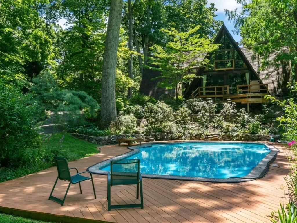 A outdoor swimming pool surrounded by a spacious wooden deck with lounge chairs, set below the main A-frame house which is nestled in a dense, mature forest.