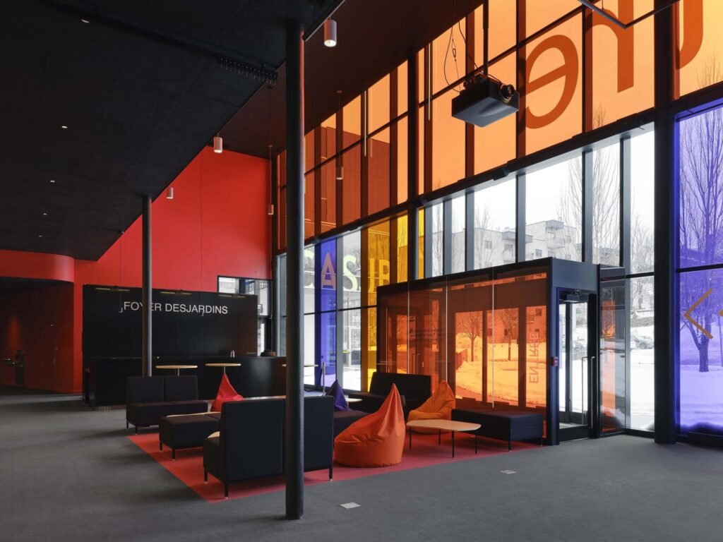 High-ceiling interior lobby of Le Grand-Espace filled with natural light passing through colorful glass panels and furnished with colorful seating.