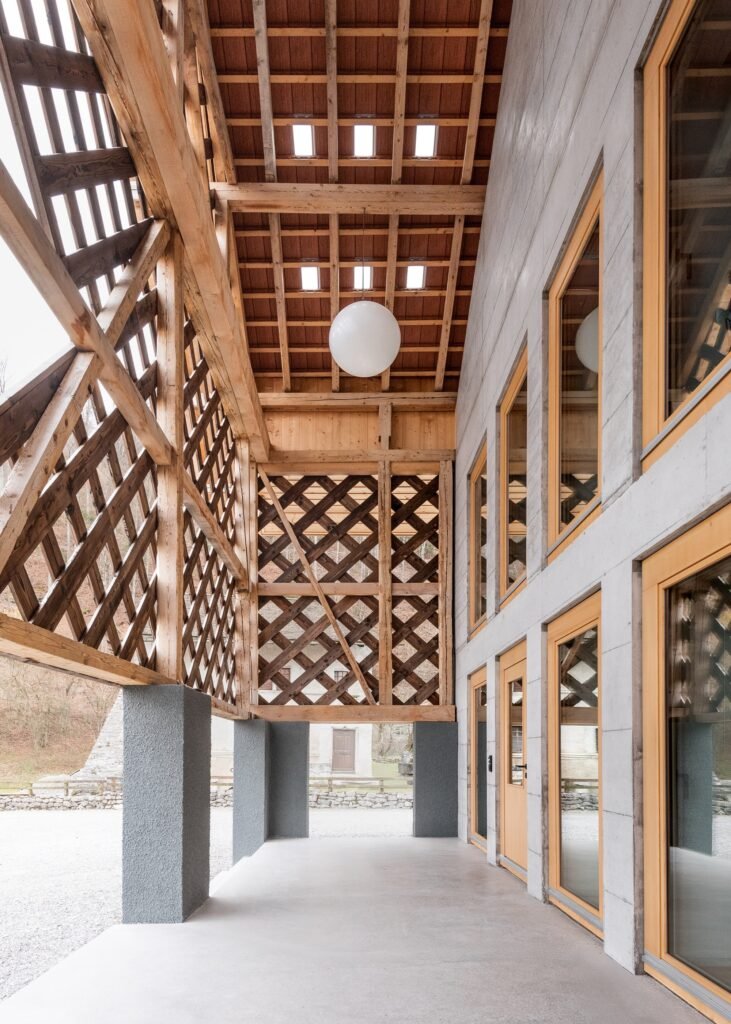 Two-story covered outdoor gallery with exposed wooden roof trusses, timber lattice walls, hanging globe light, and full-height glass doors along a concrete wall.
