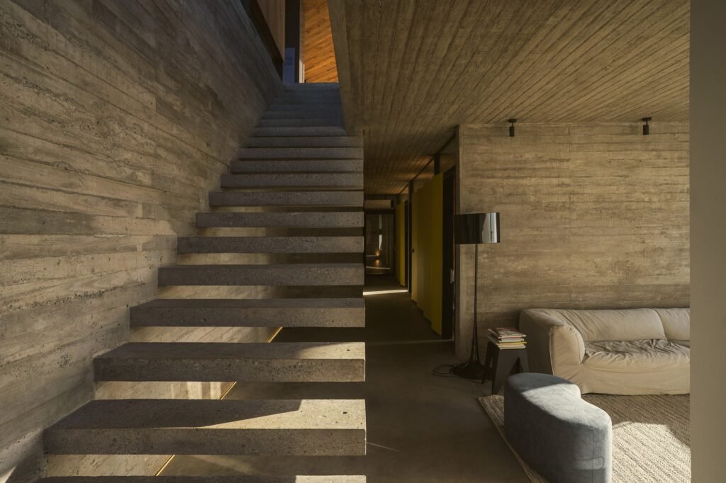 Interior of Casa 22 displaying board-formed concrete walls, a floating concrete staircase, warm wooden ceilings, and soft indirect lighting.