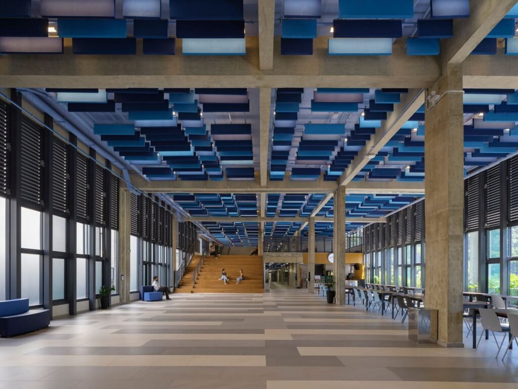 Spacious internal atrium inside Colégio Bernoulli featuring blue acoustic panels mounted on a concrete ceiling grid and wide stairs.
