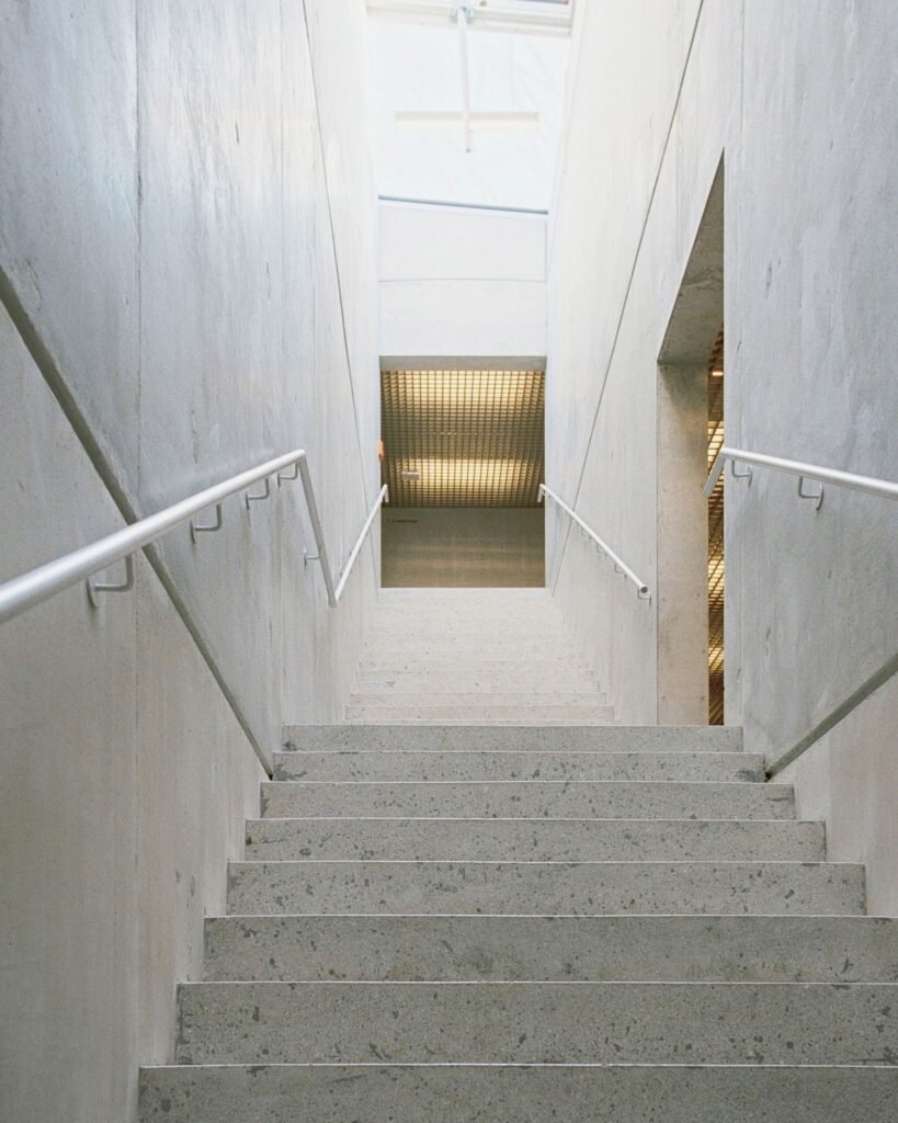 Straight concrete staircase with smooth white metal handrails flanked by fair-faced concrete core walls under overhead natural daylight.