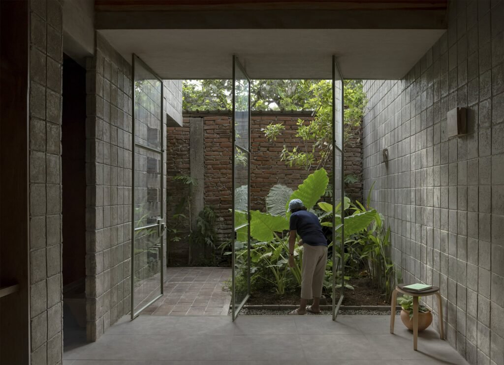 Inside La Colmena, a resident tends to lush green plants in one of the open-air filtration patios, seen through open, pivot glass-and-steel doors from a concrete-block-walled interior.