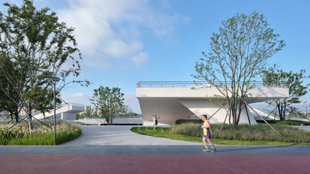 A jogger running along a red track past a contemporary white concrete pavilion with an elevated viewing deck under a clear blue sky.