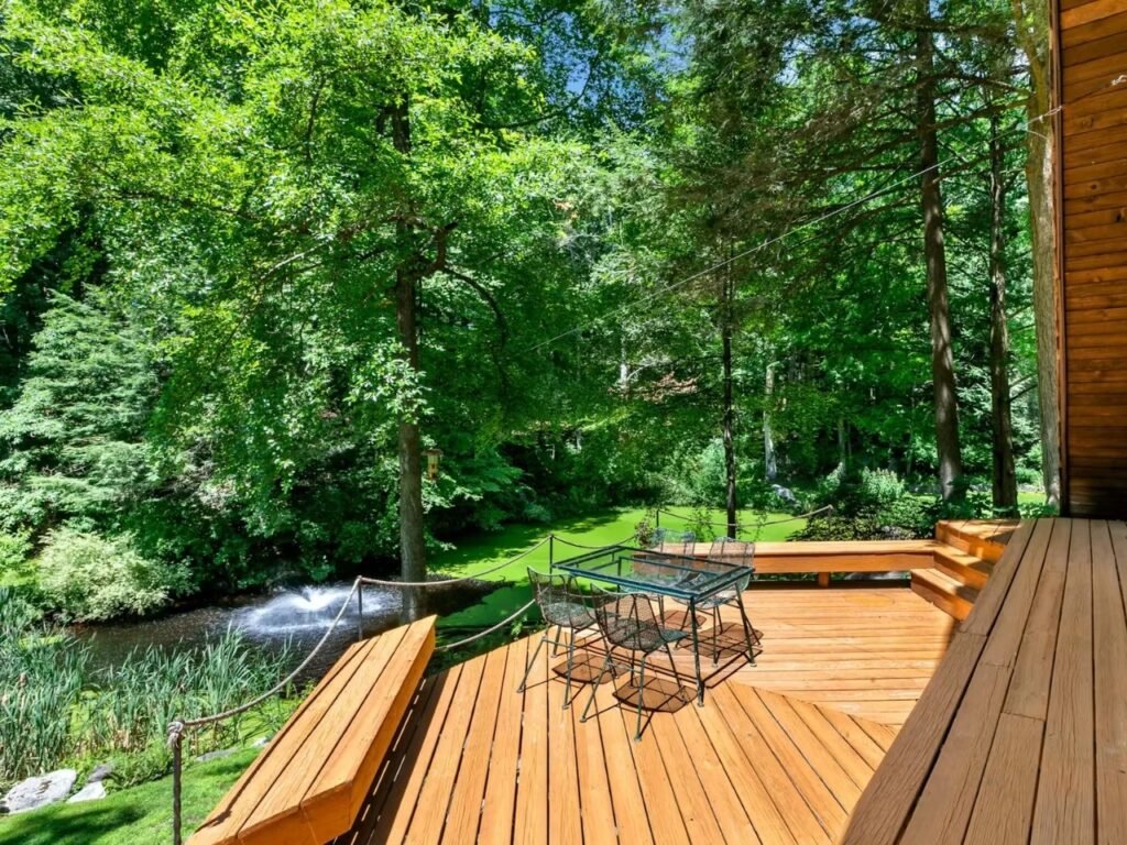 A view from an elevated wooden deck equipped with built-in bench seating and a patio dining table, looking out over a natural pond with a water fountain surrounded by trees.