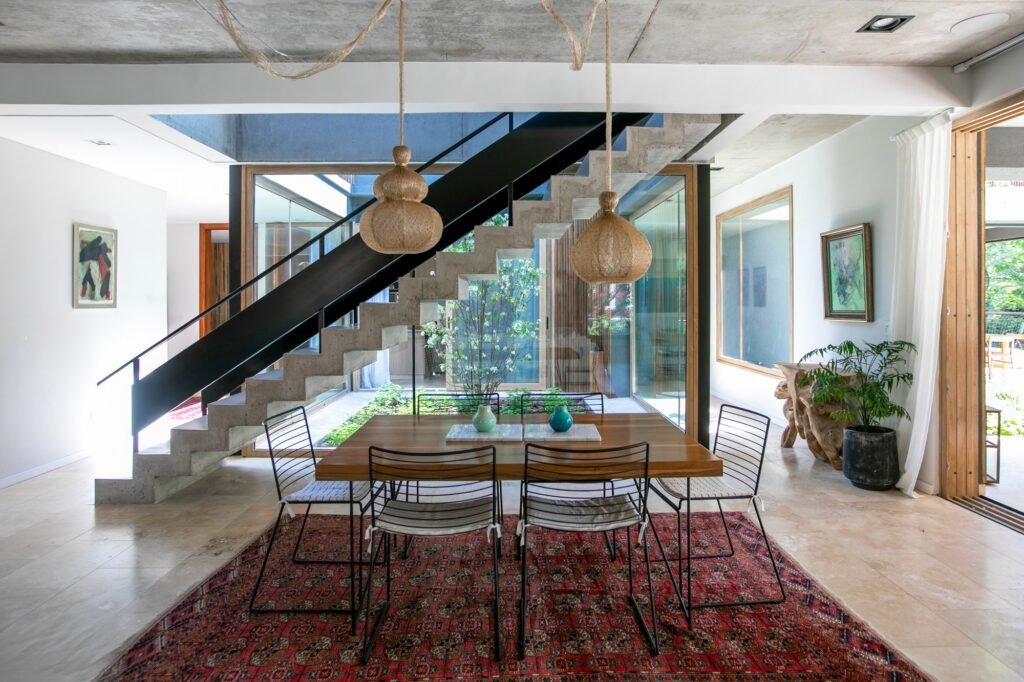 Modern dining room with a wooden table, concrete staircase, woven pendant lights, and floor-to-ceiling glass looking onto an internal courtyard.