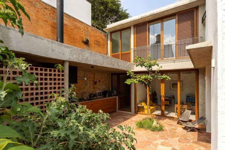 Outdoor courtyard with sandstone paving, a central young tree, cobogo ceramic brick wall, and covered barbecue area in Casa em São Paulo.