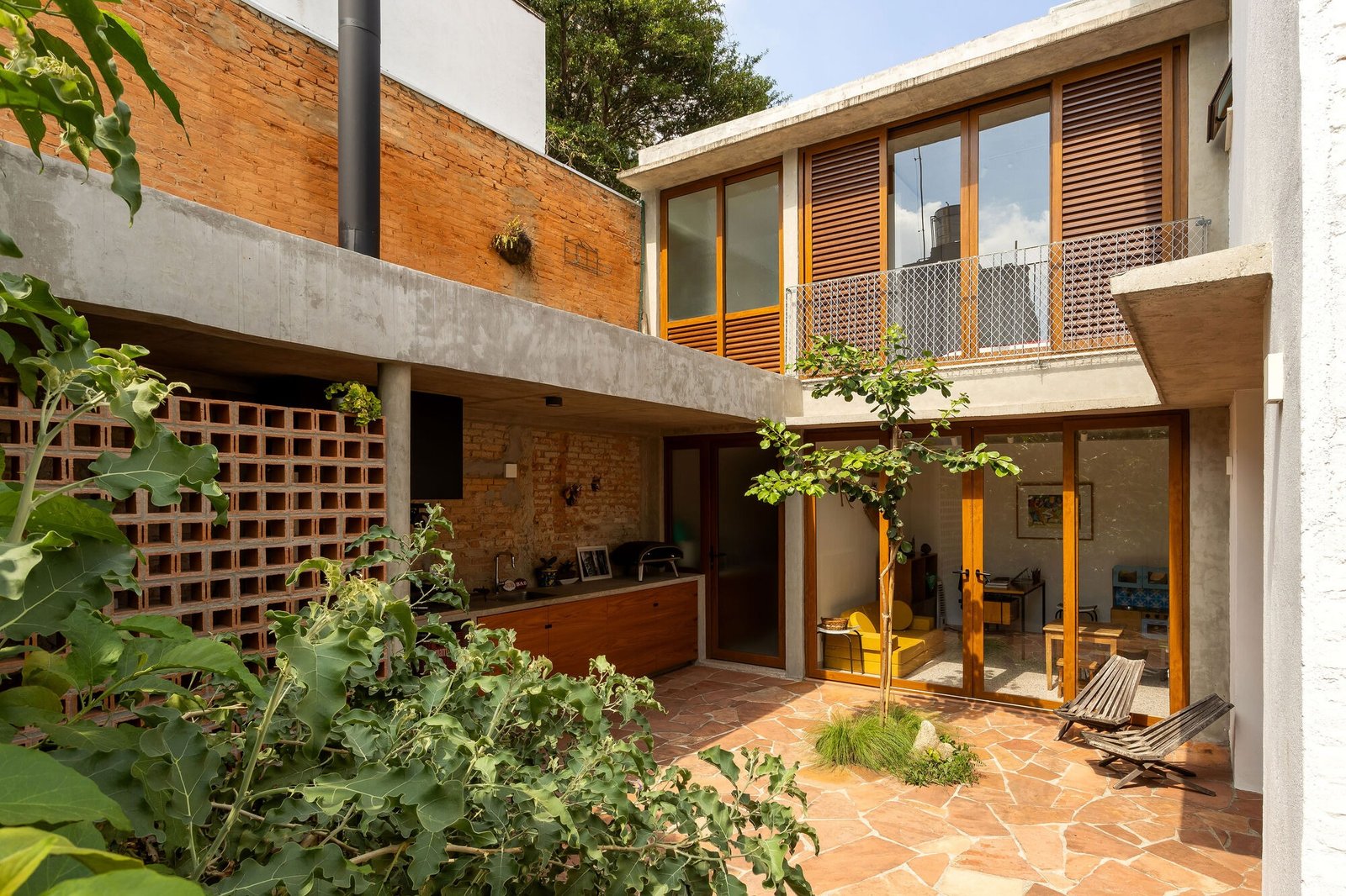 Outdoor courtyard with sandstone paving, a central young tree, cobogo ceramic brick wall, and covered barbecue area in Casa em São Paulo.