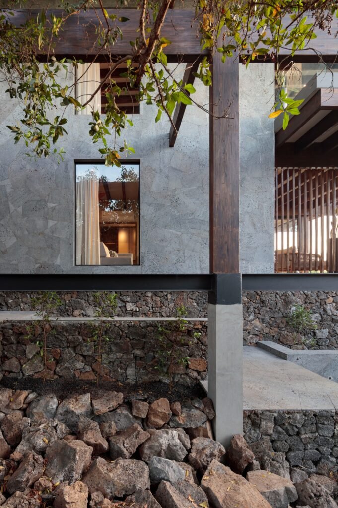Close-up of a facade section of the Finch Bay Hotel, showing textured lava stone walls, dark laminated wood beams, glass-railed balconies, and a metal beam base. Plants sprout from rocky ground, with the illuminated room visible through a window.
