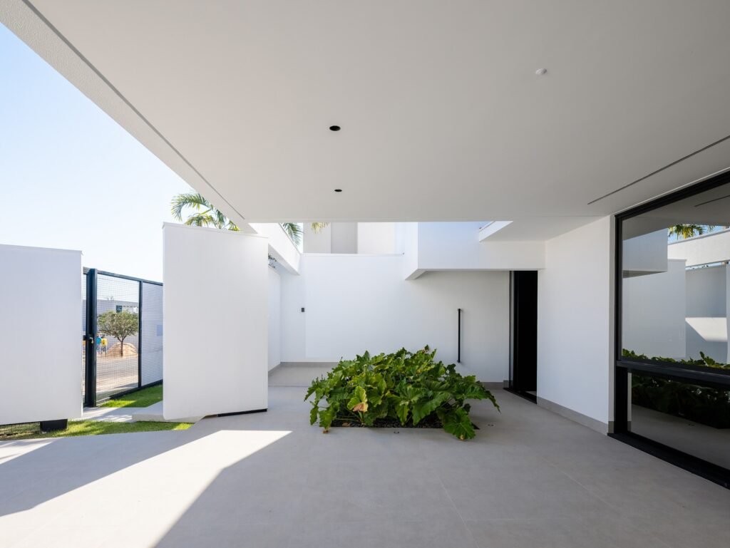View from a covered outdoor patio or garage area at Sorriso House, looking toward an open-air courtyard with a central planter, white walls, and an industrial-style gate.