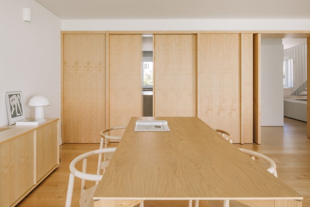 Minimalist interior dining space featuring a light wooden table, chairs, and integrated wooden wall paneling with sliding doors.