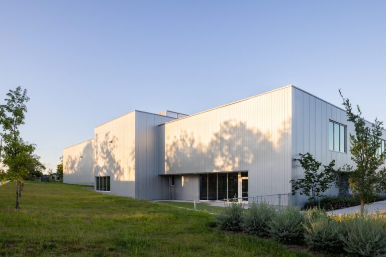 Metallic corrugated metal facade of the Crux South building framed by green lawn and trees.